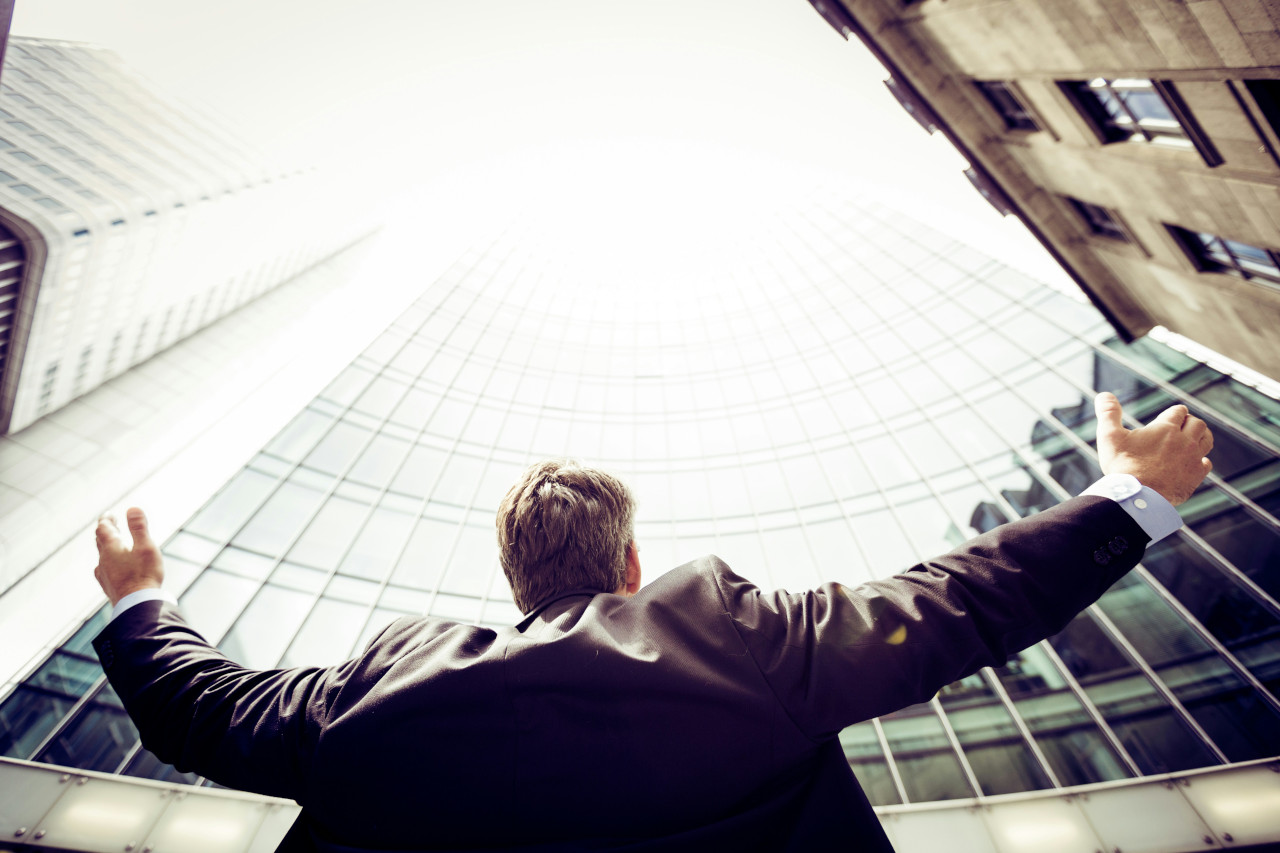 A confident business professional in a suit stands with arms outstretched, gazing upwards toward a towering glass building, symbolizing achievement, growth, and the transformative power of expert guidance.